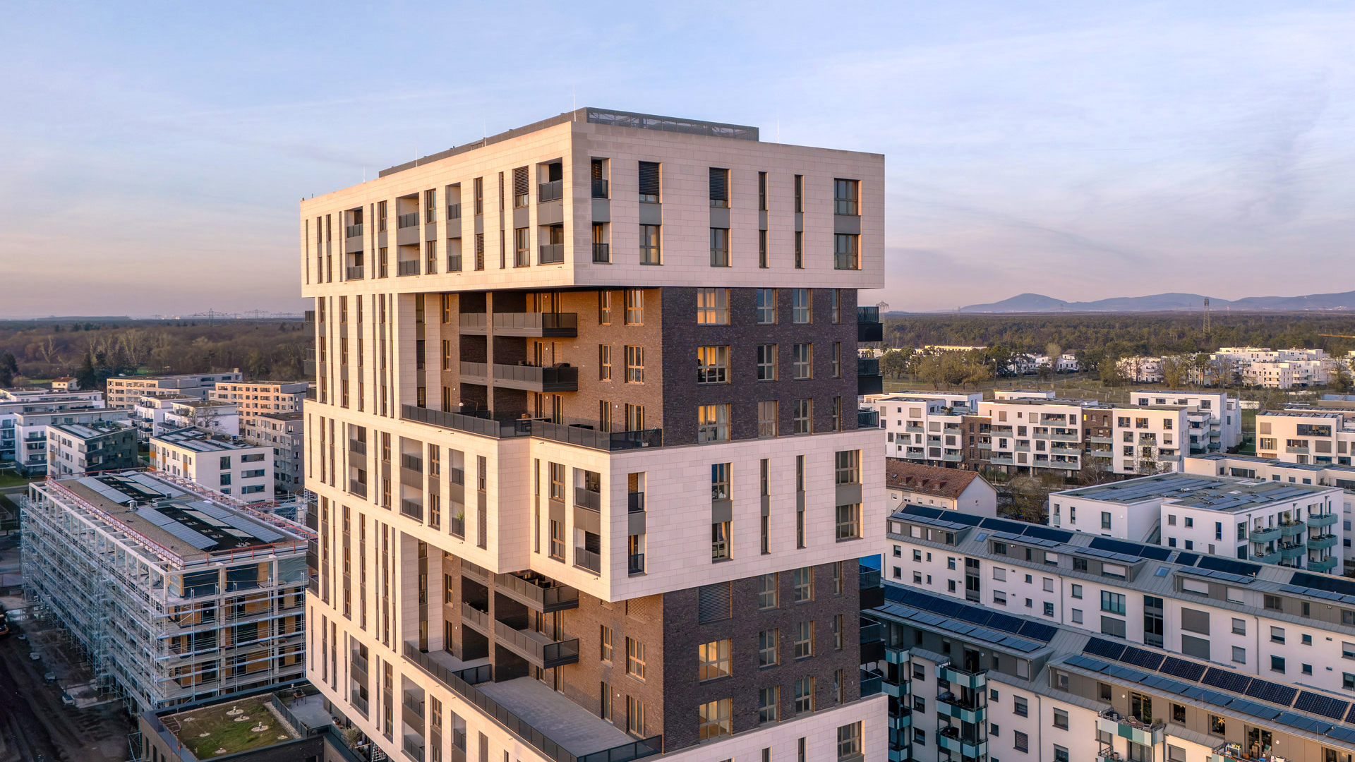 KI-basierte Bildbeschreibung: Ein modernes Wohnhochhaus mit hellen und dunklen Fassadenabschnitten steht bei Sonnenuntergang zwischen niedrigeren Wohngebäuden, im Hintergrund sind Berge und Bäume zu sehen.