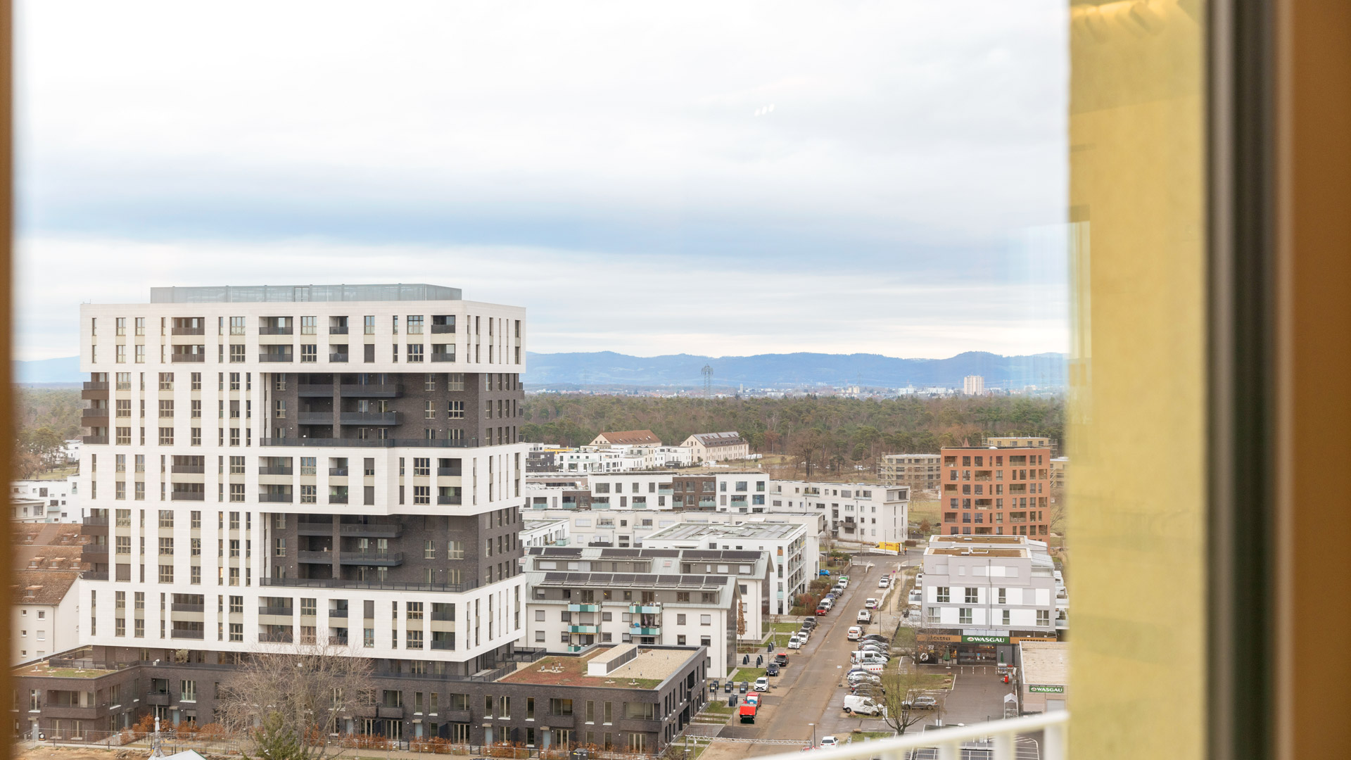 KI-basierte Bildbeschreibung: Blick auf eine moderne Stadtlandschaft mit einem hohen weiß-schwarzen Wohnhaus, kleineren Gebäuden und einer Straße mit geparkten Autos. Im Hintergrund sind Berge und ein bewölkter Himmel zu sehen. Rechts ist ein Teil eines Fensterrahmens zu sehen.