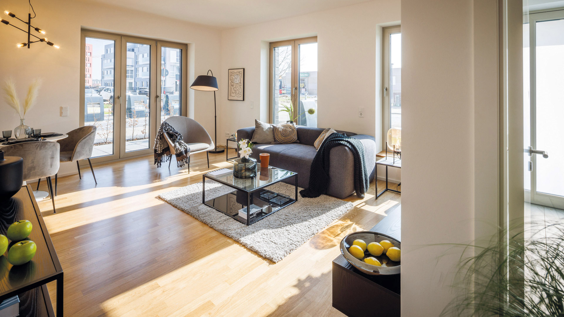 KI-basierte Bildbeschreibung: Ein modernes, sonnendurchflutetes Wohnzimmer mit großen Fenstern, einem grauen Sofa, einem Couchtisch aus Glas, einem Sessel und einem Holzboden. Eine Essecke mit Stühlen ist zu sehen, zusammen mit dekorativen Pflanzen und Schalen mit Äpfeln und Zitronen.