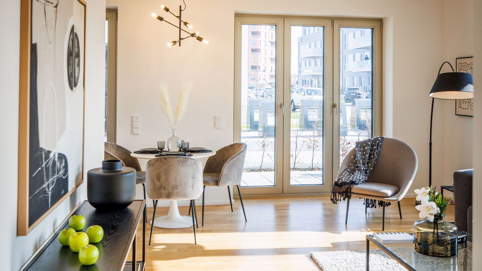KI-basierte Bildbeschreibung: Ein moderner, heller Wohn- und Essbereich mit hellem Holzfußboden, rundem Esstisch mit Samtstühlen, großen Fenstern und schlichter, zeitgenössischer Einrichtung mit Kunstwerken, einer Stehlampe und einem kleinen Couchtisch mit Blumen.