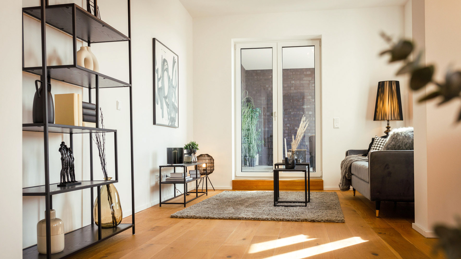 KI-basierte Bildbeschreibung: Ein modernes Wohnzimmer mit Holzböden, minimalistischen Möbeln, einem grauen Sofa, einer Stehlampe, einem Teppich und dekorativen Regalen. Große Glastüren lassen natürliches Licht herein und schaffen einen hellen, gemütlichen Raum.