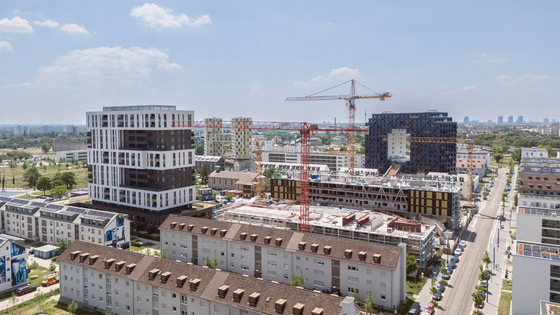 KI-basierte Bildbeschreibung: Eine Stadtlandschaft zeigt moderne Wohngebäude und Baukräne, die an neuen Hochhäusern arbeiten, unter einem blauen Himmel mit vereinzelten Wolken. Im Hintergrund sind Wohnstraßen und Grünflächen zu sehen.